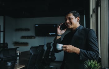 A man wearing a blazer holds a cup of coffee while speaking on a cell phone.